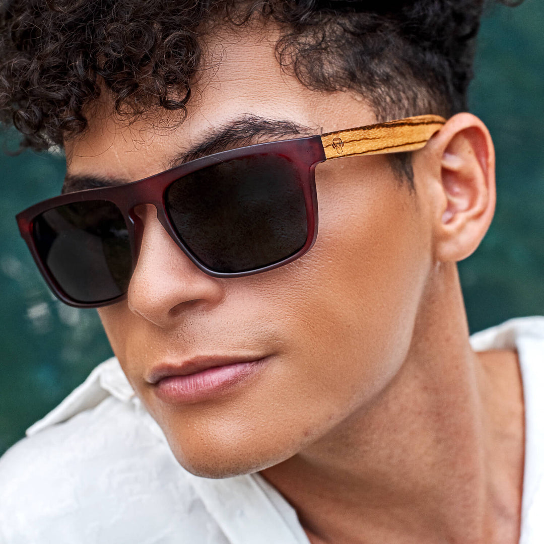 Un modelo pelo rulos in primer plano mirando in 45 grados to the left wearing sunglasses red in recycled plastic with wooden temples natural Bonoboss #color_wine