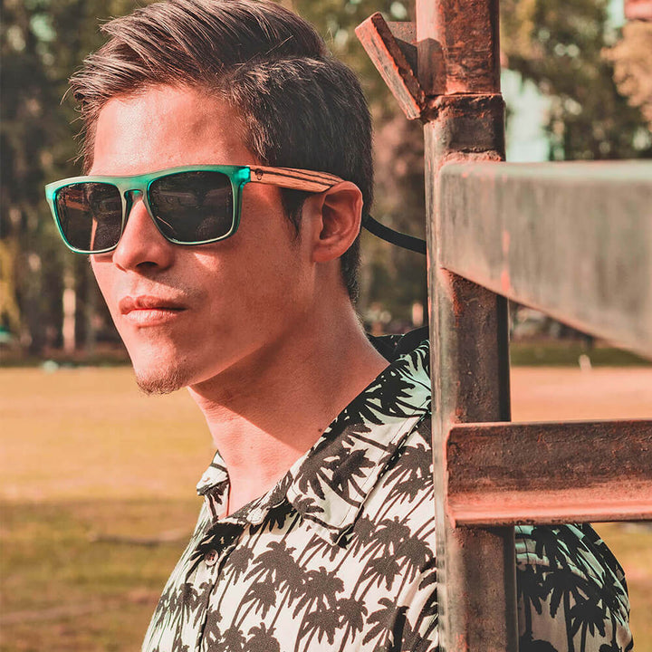Un men serio in 45 grados with camisa blanca and a pattern palmeras black wearing sunglasses polarized green hecho with recycled plastic and wooden temples natural Bonoboss #color_forest