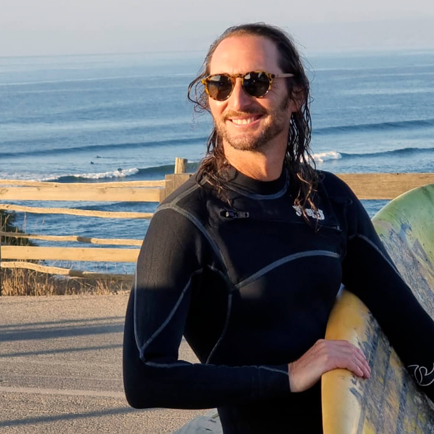 un modelo hombre en la playa con traje de surf y sosteniendo una tabla de surf usando lentes hexagonales color carey con lentes polarizadas. El hombre está sonriendo y de fondo se ve el mar de la playa pichilemu #color-marco_carey